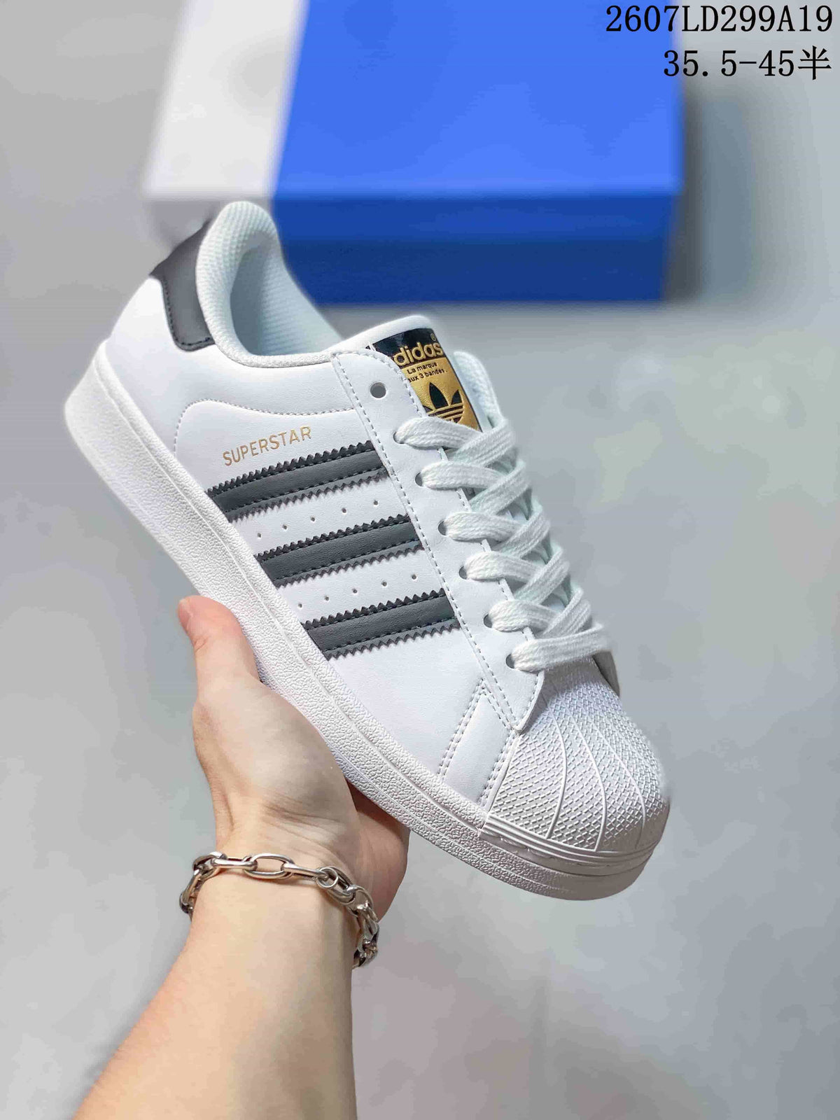 White adidas Superstar sneaker with dark gray serrated stripes, gold side branding, and a gold-and-black tongue label.