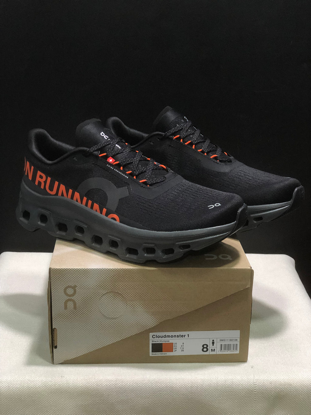 Pair of black and orange On Cloudmonster 1 sneakers displayed on a branded shoe box; the box label shows a photographed size of US 8, EU 41, UK 7.5, and JP 26.