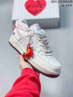 Angled side view showing the white sneaker, pale pink Swoosh, burgundy outsole, and attached decorative charm.