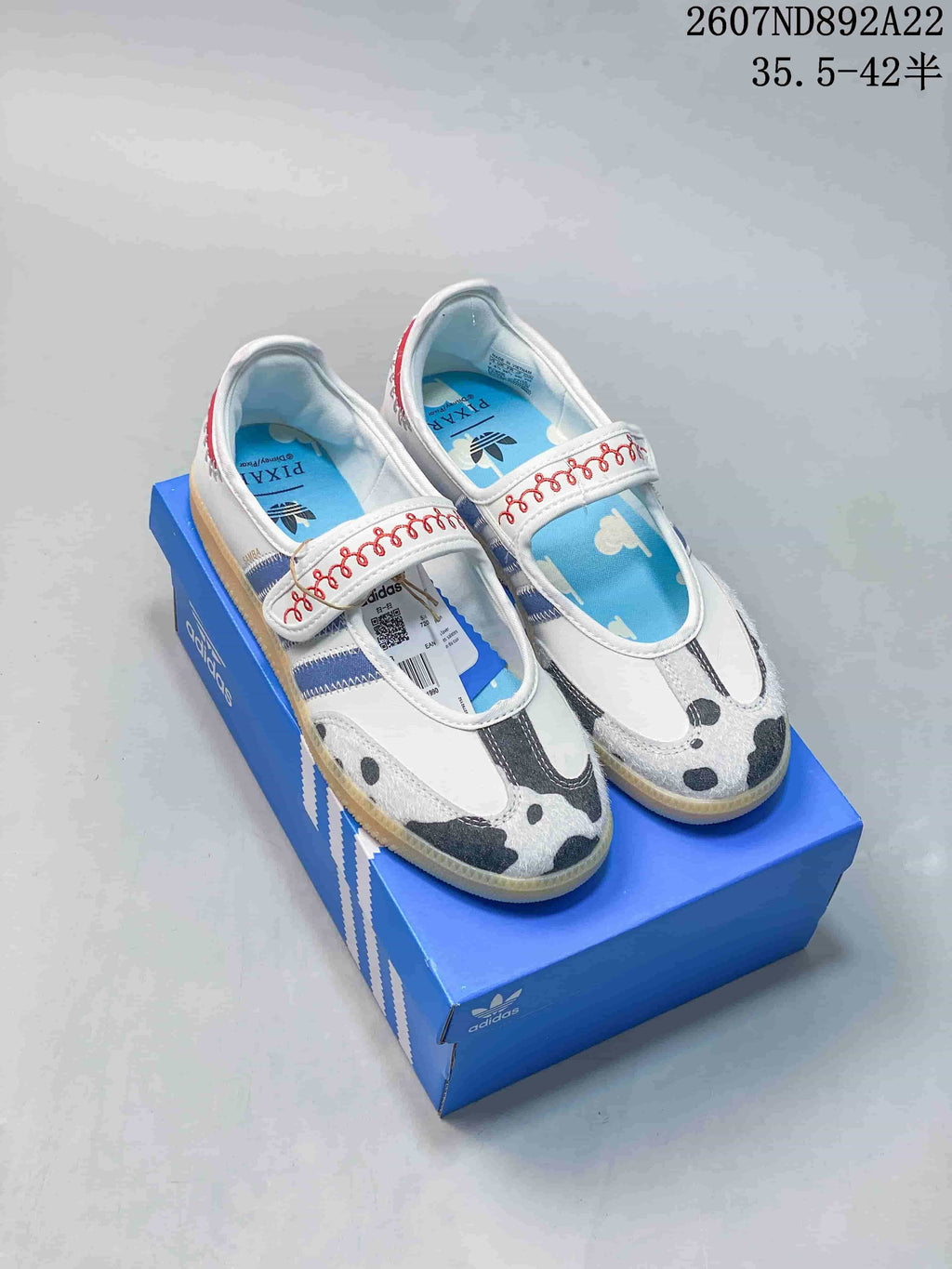 Pair of white adidas Samba Jane sneakers with blue stripes, red strap detailing, patterned toe panels, and a blue shoebox.