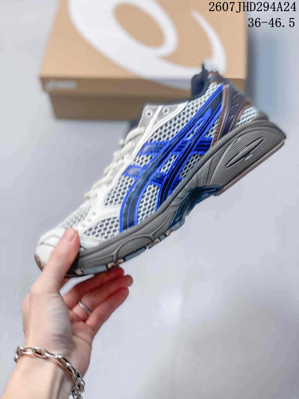 Side profile of the ASICS sneaker showing the mesh-style upper, layered overlays, blue side branding, and gray midsole.