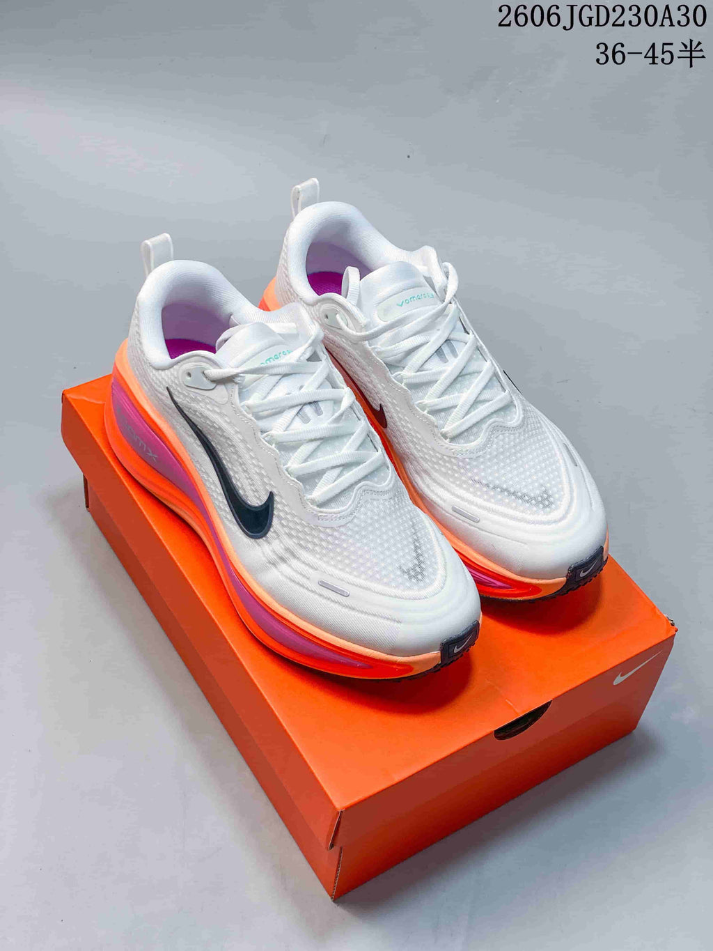 Pair of white Nike Vomero Plus shoes with dark Swooshes and orange and pink midsole accents, on an orange shoebox.