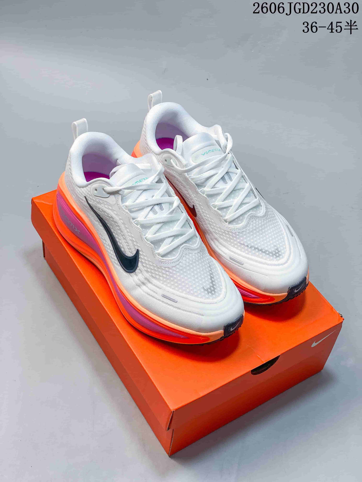 Pair of white Nike Vomero Plus shoes with dark Swooshes and orange and pink midsole accents, on an orange shoebox.