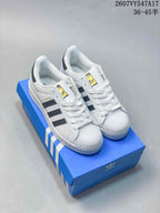 Pair of white Superstar sneakers displayed on a blue adidas shoebox, with Trefoil tongue branding visible.