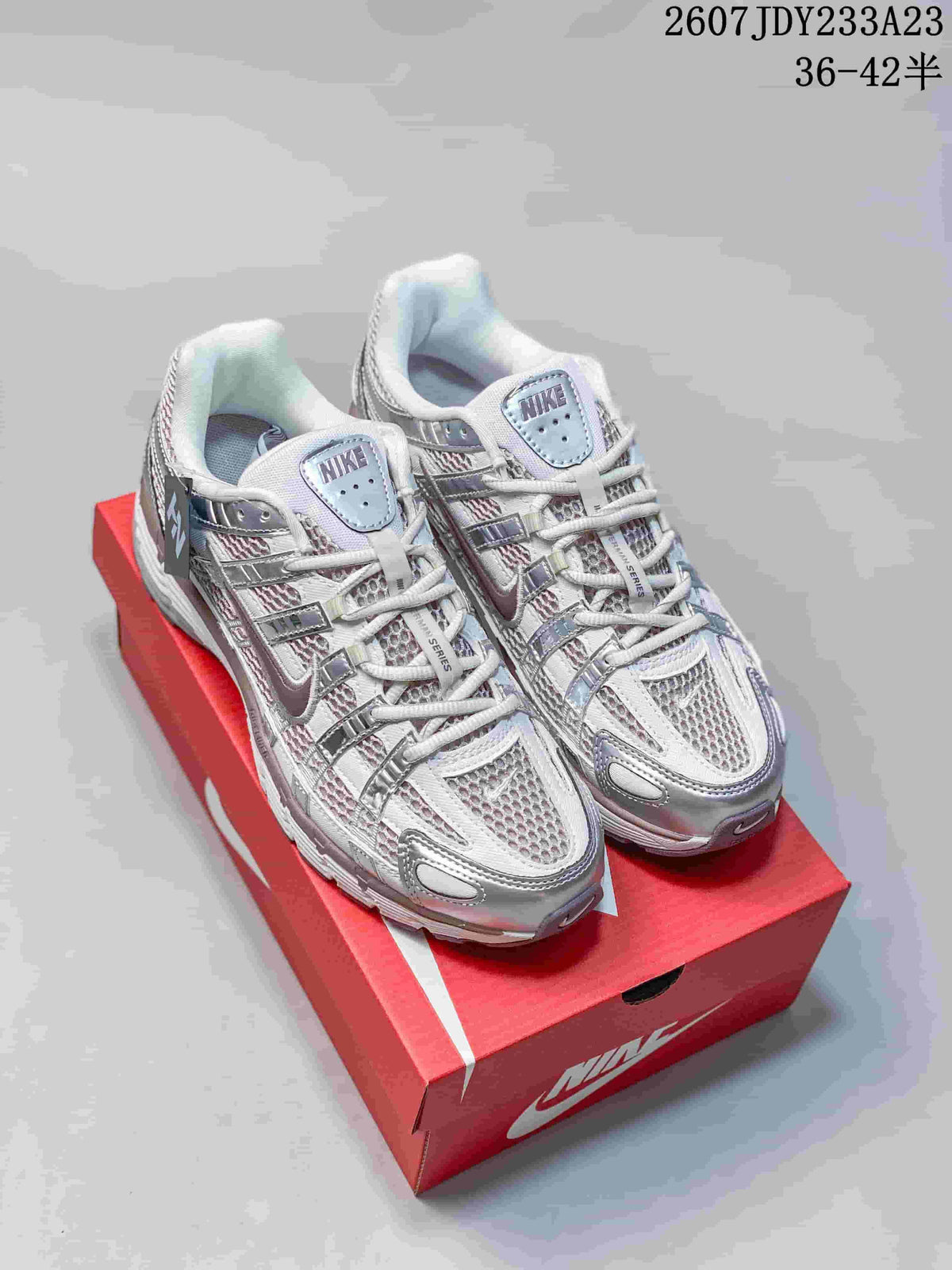 Front three-quarter view of white and silver Nike P-6000 sneakers with red and gray accents on a red shoebox; the image shows style code 2607JDY233A23 and size range 36–42.