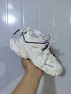 White Balenciaga sneaker held at an angle, showing the layered upper, chunky outsole, and navy and lavender laces.