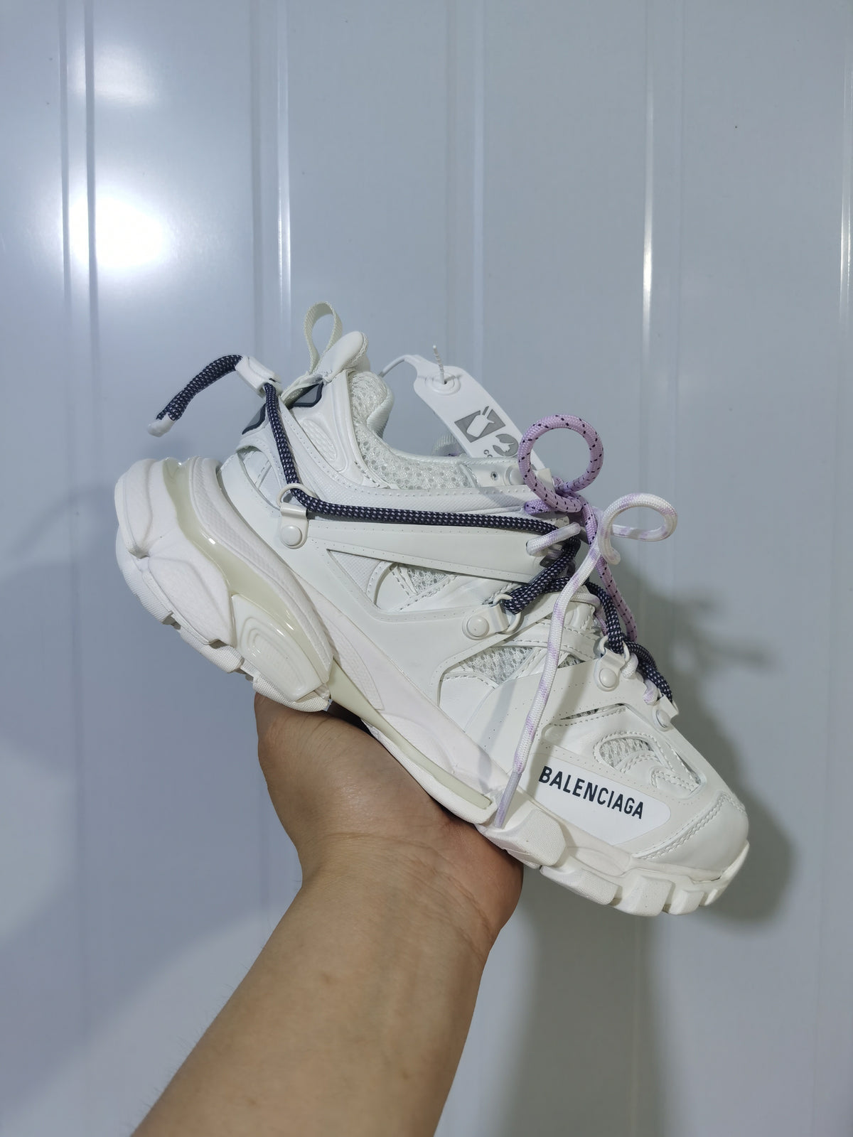 White Balenciaga sneaker held at an angle, showing the layered upper, chunky outsole, and navy and lavender laces.