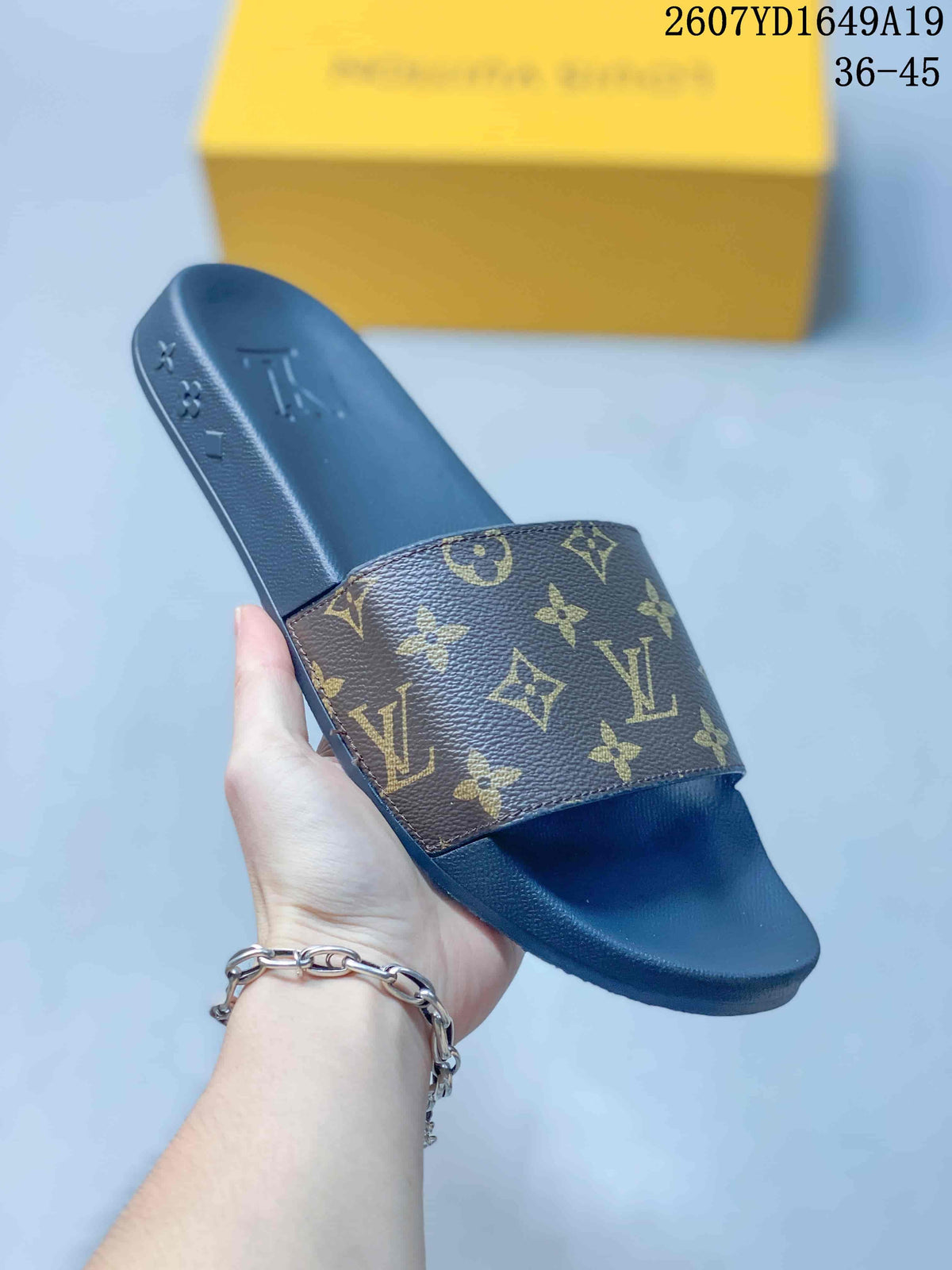 Close view of a brown-and-gold monogram slide sandal held by hand, showing the wide upper and dark platform-style sole.