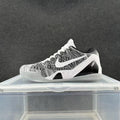 Gray and black Nike low-top sneaker with a textured upper, white Swoosh, white laces, and contrasting midsole displayed on a clear stand.