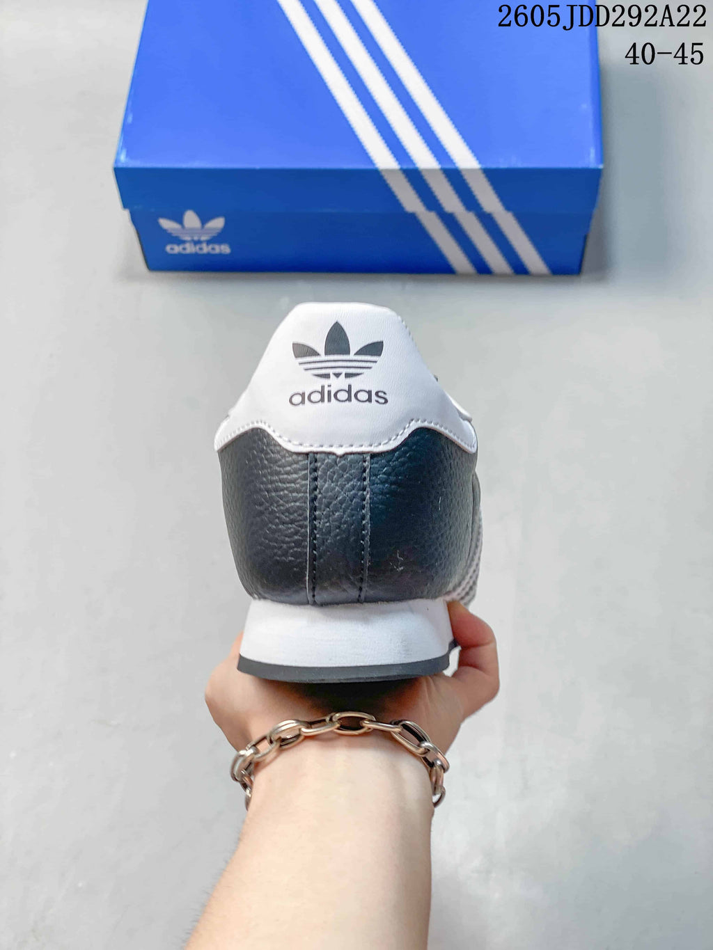 Rear view of a dark gray adidas Samoa sneaker with a white heel accent and blue shoebox in the background.
