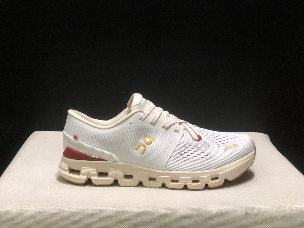 Side profile of a white On Cloud X4 sneaker with gold branding, burgundy heel detail, and cream-colored outsole.