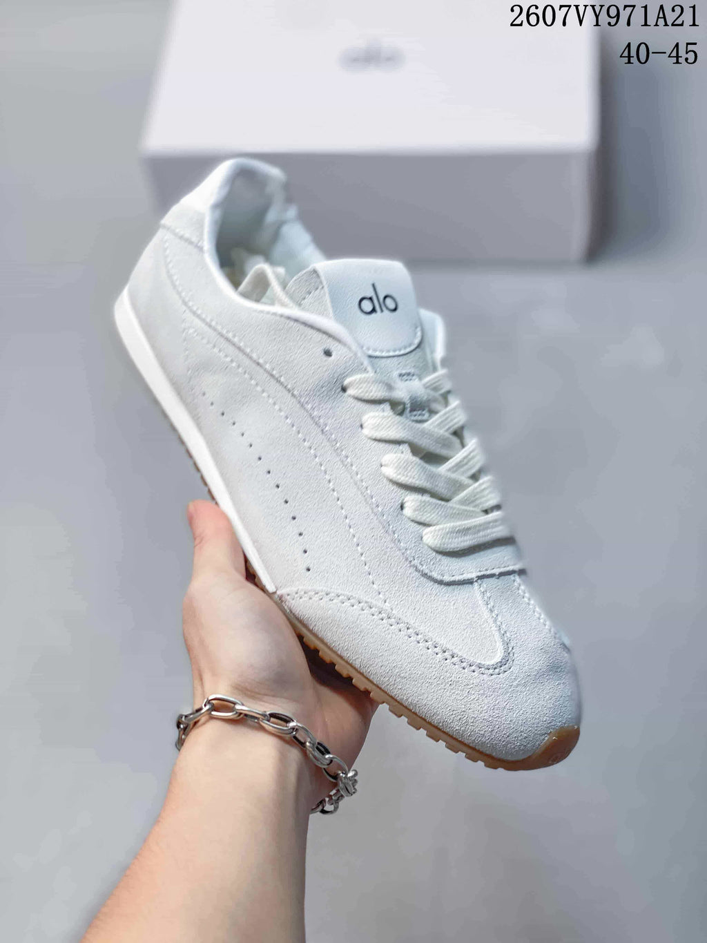Close side view of a white Alo Yoga sneaker showing layered panels, perforated detailing, white laces, and a gum-colored outsole.