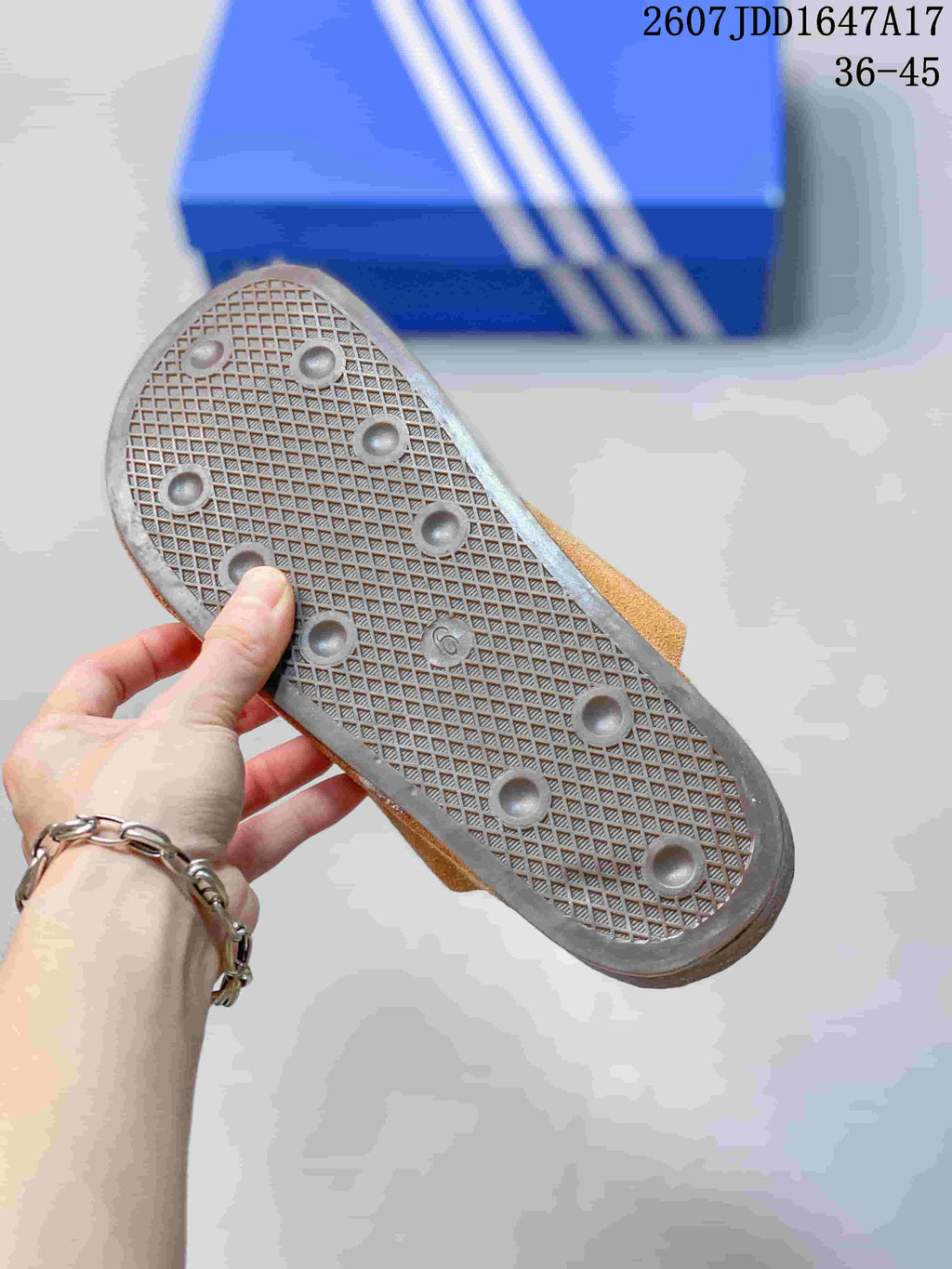 Underside of a tan slip-on mule showing a textured crosshatch outsole with circular traction elements and a blue adidas shoebox in the background.