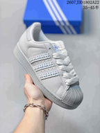 Close-up side view of a white and gray Adidas Superstar sneaker showing the shell-style toe, white laces, and decorative side stripes.