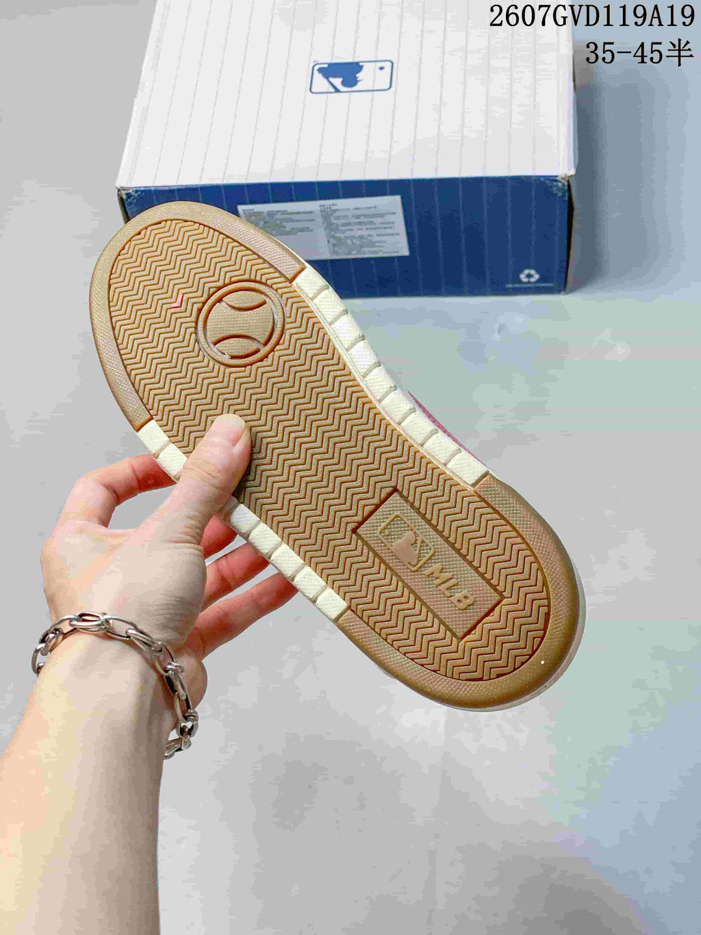 Outsole view of a sneaker showing a textured herringbone tread pattern, gum-colored base and MLB branding, with the product box in the background.