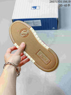 Outsole view of a sneaker showing a textured herringbone tread pattern, gum-colored base and MLB branding, with the product box in the background.