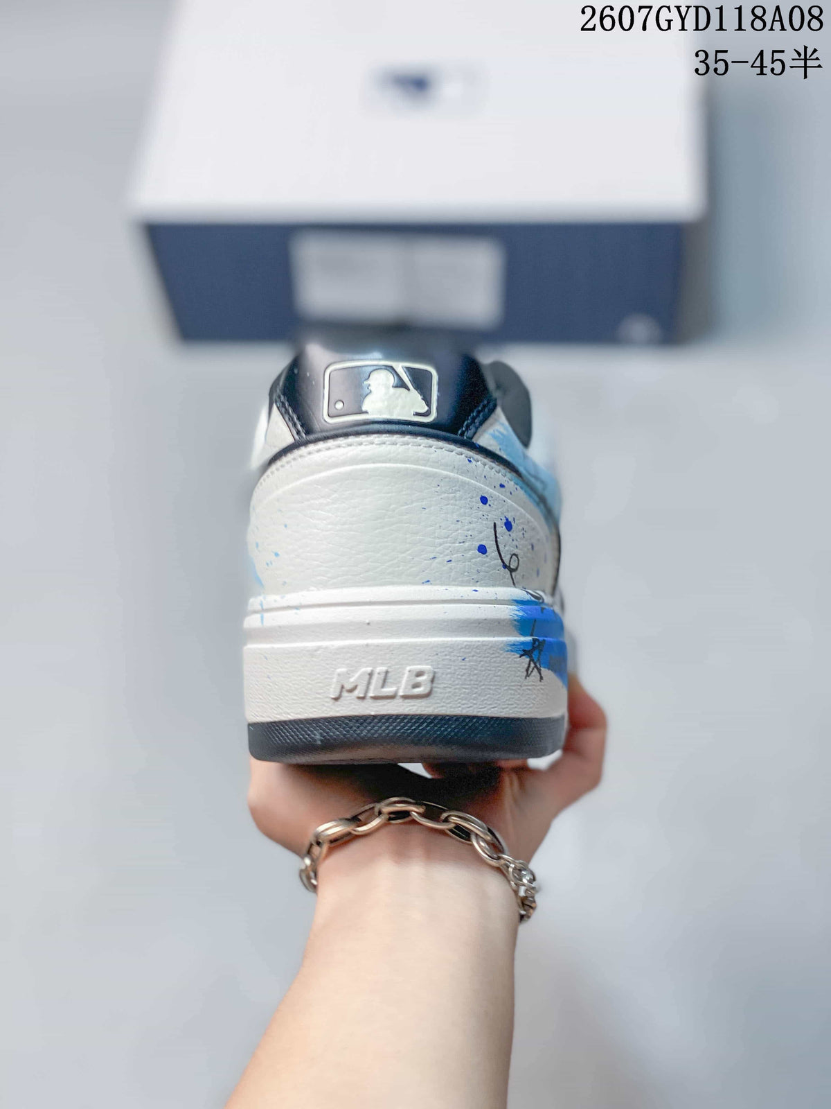 Rear view of the white sneaker showing blue splatter details, an MLB-marked heel section and dark outsole trim.