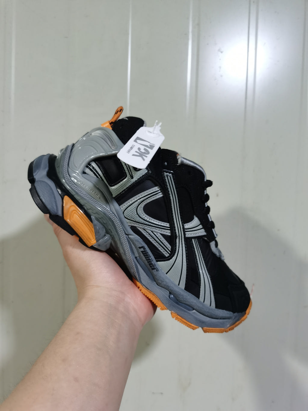 Top and side view of the sneaker showing black laces, grey structural overlays, orange outsole accents and a hanging brand tag.