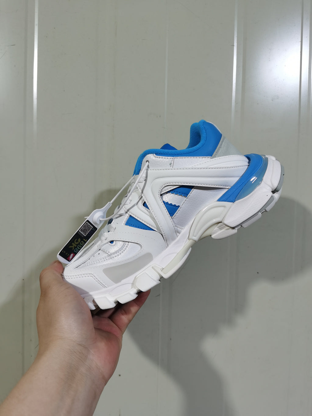 Reverse angled view of the white and blue low-top sneaker showing the layered upper, chunky outsole, heel detail, and attached product tag.