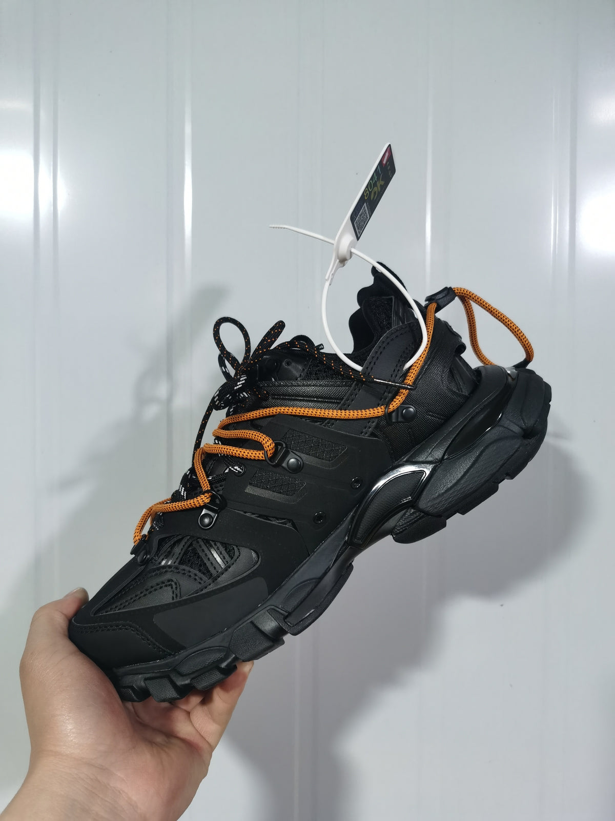 Angled view of a black Balenciaga Track sneaker with layered panels, orange and black rope-style laces, and a rugged segmented outsole.