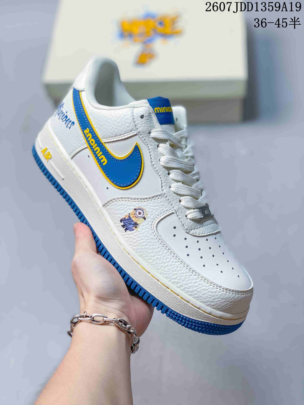 Close-up of a white low-top sneaker with a blue-and-yellow Swoosh, white laces, blue outsole, and small character artwork; image text shows style code 2607JDD1359A19 and size range 36-45.