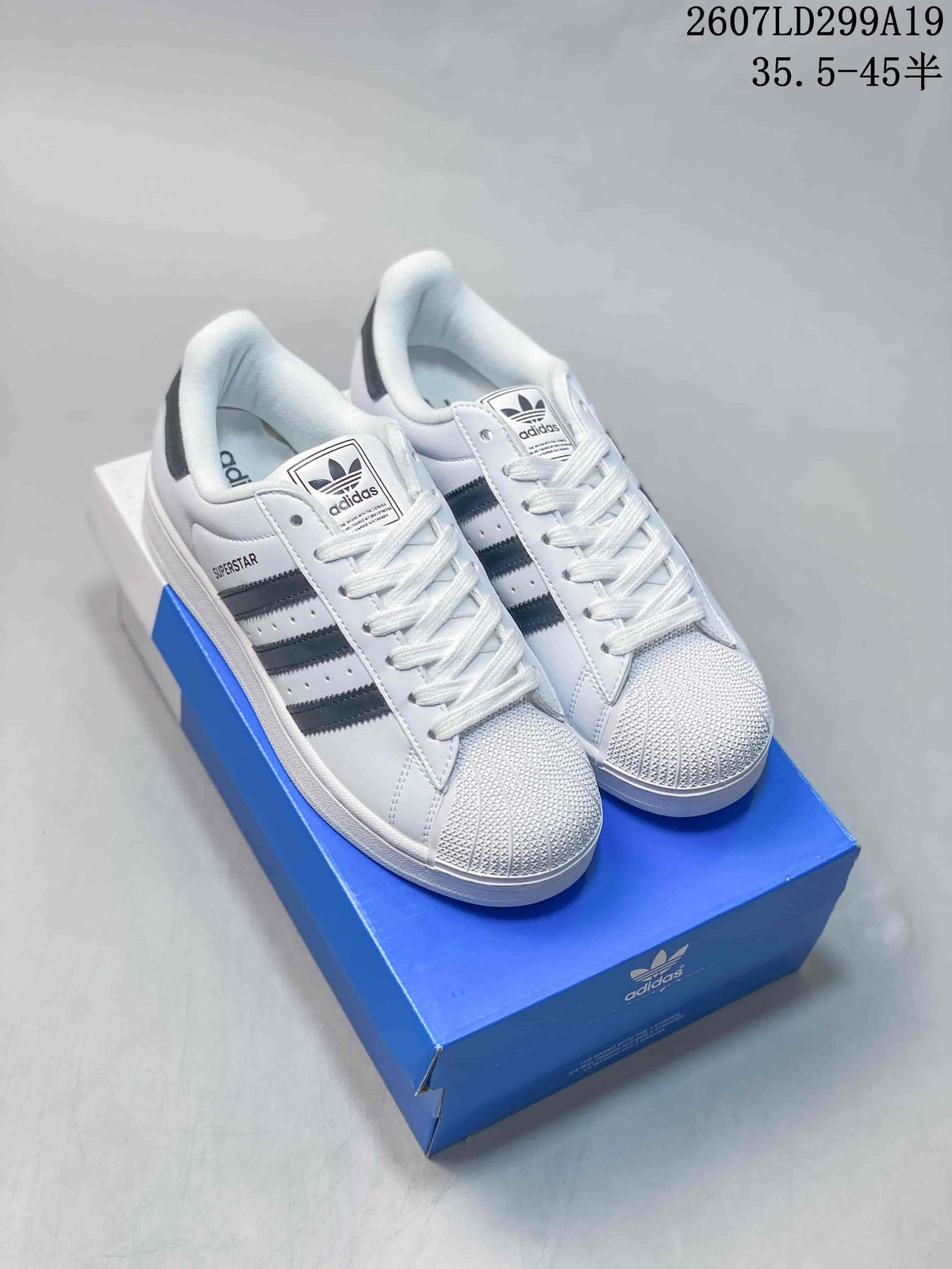 Pair of white adidas Superstar sneakers with black stripes and shell toes displayed on a blue adidas shoe box; image text shows style code 2607LD299A19 and size range 35.5–45 half sizes.