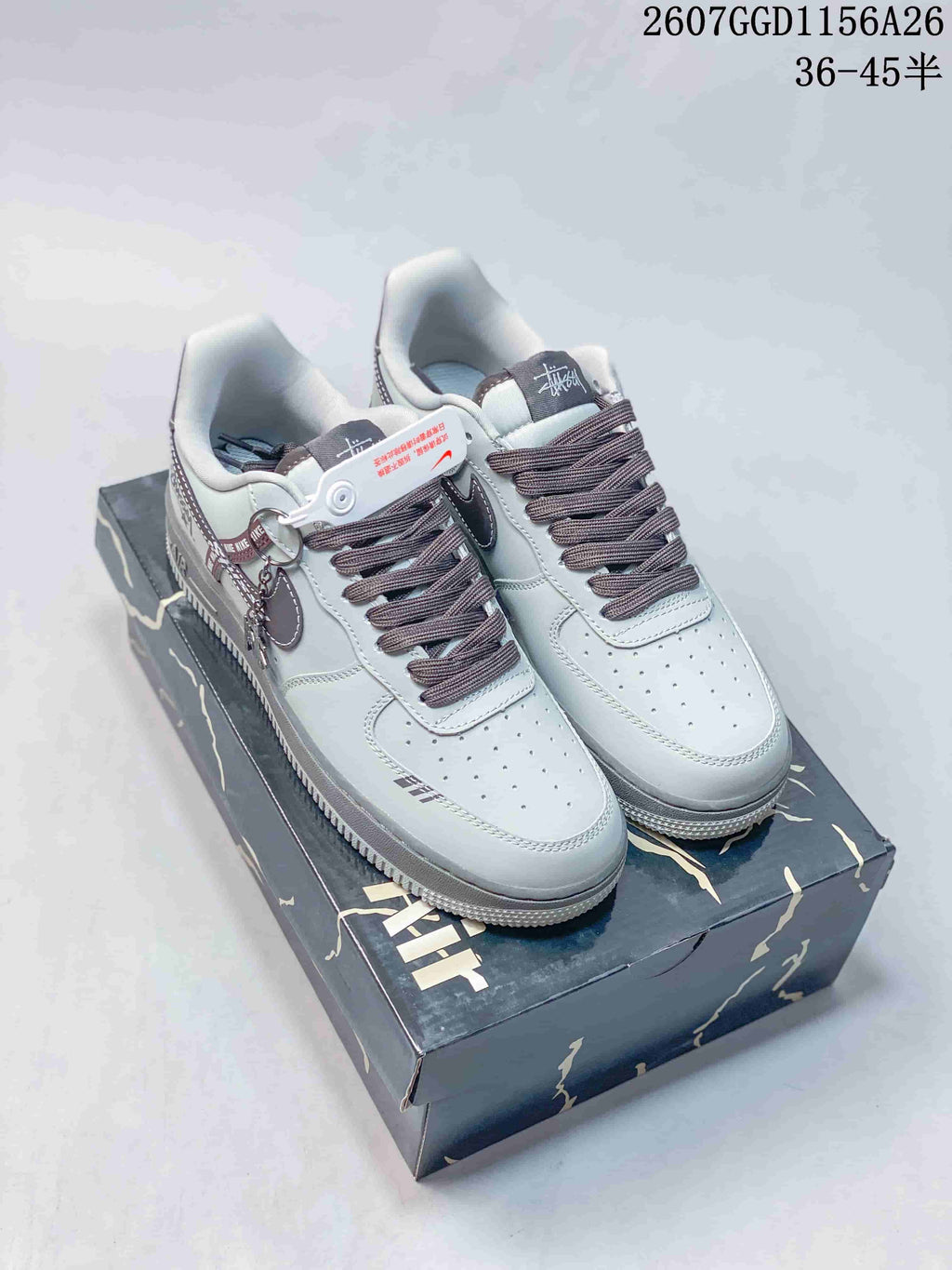 Pair of gray low-top Nike Air Force 1 sneakers displayed on a patterned shoe box; overlay text shows style code 2607GGD1156A26 and sizes 36–45.