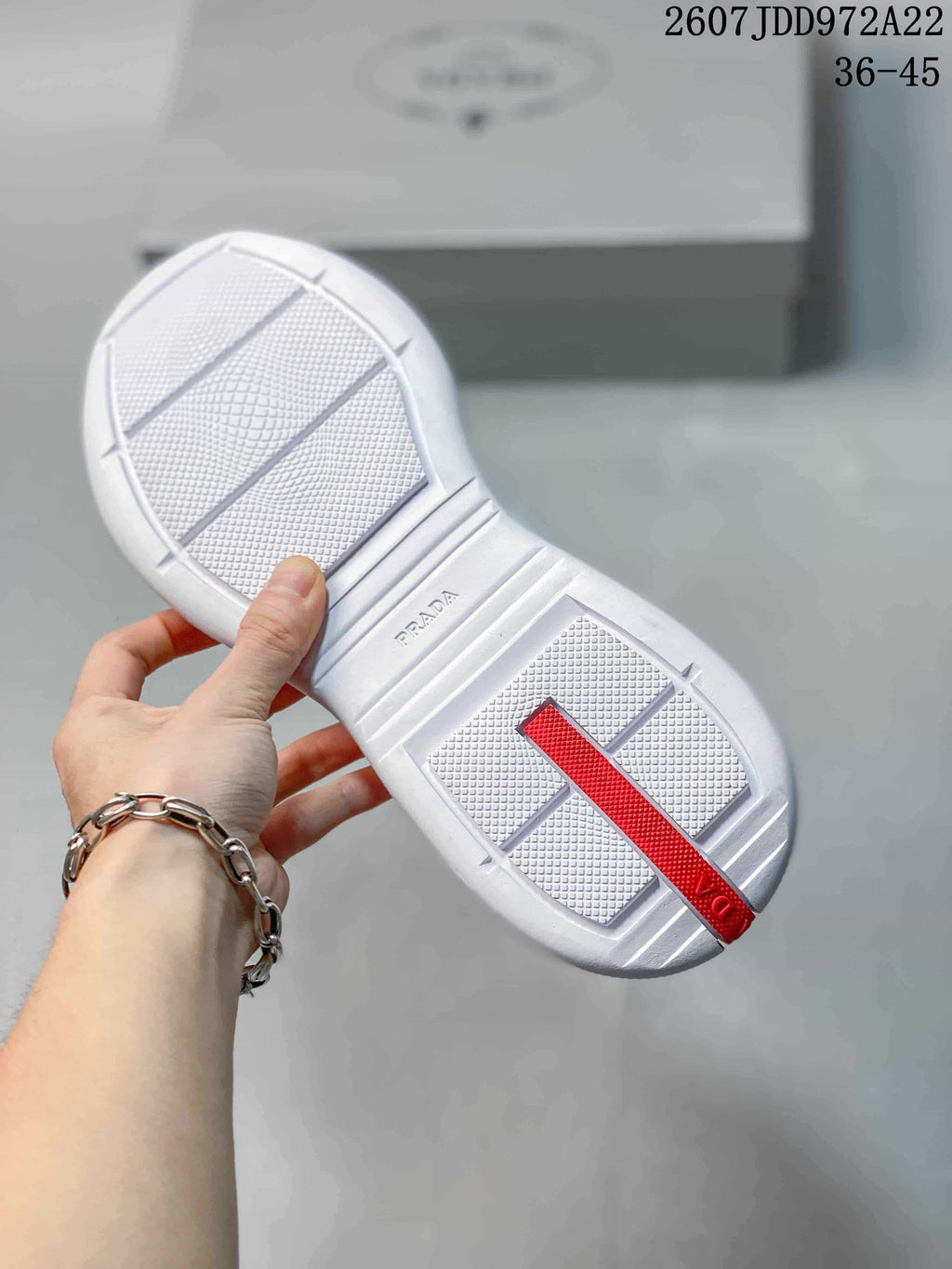 Underside view of a white sneaker showing the textured outsole, central branding detail, and red heel strip.