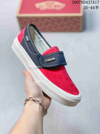 Side view of a red and navy Vans slip-on sneaker showing the strap, suede-textured upper, Vans branding, and white rubber sole.