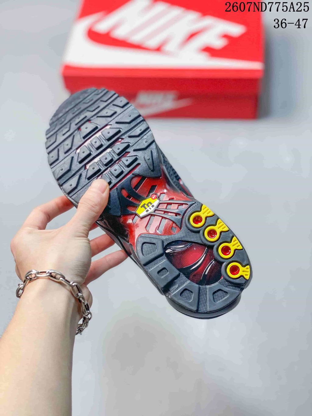 Underside view of a black Nike Air Max Plus TN sneaker showing the patterned outsole with yellow and red details; overlay text shows code 2607ND775A25 and size range 36–47.