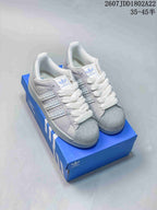 Pair of pale gray and white Adidas Superstar sneakers shown from a front three-quarter angle on a blue shoe box.
