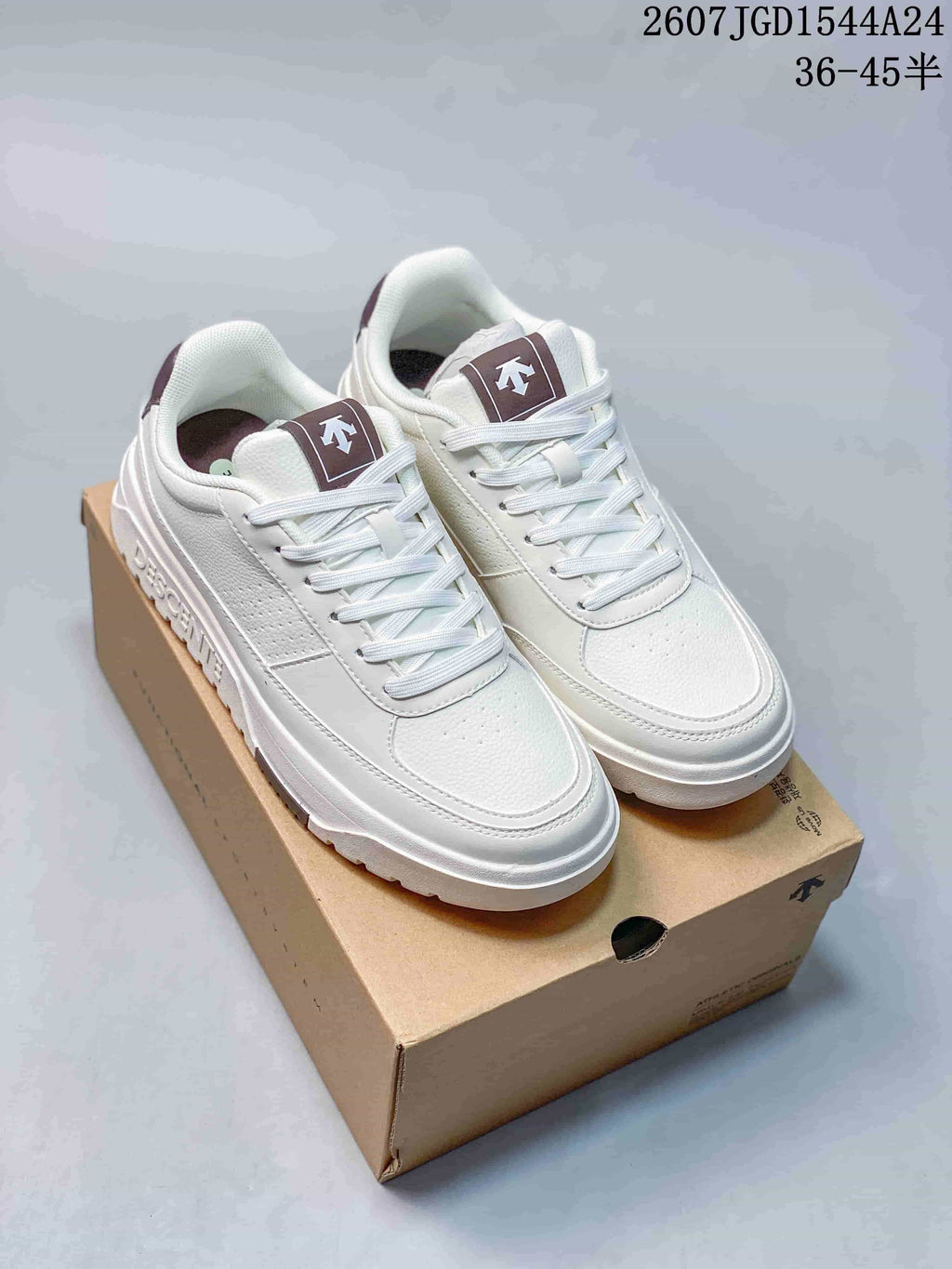 Pair of white DESCENTE Dualis low-top sneakers with brown tongue accents displayed on a shoe box; overlay shows style code 2607JGD1544A24 and size range 36–45 with half sizes.