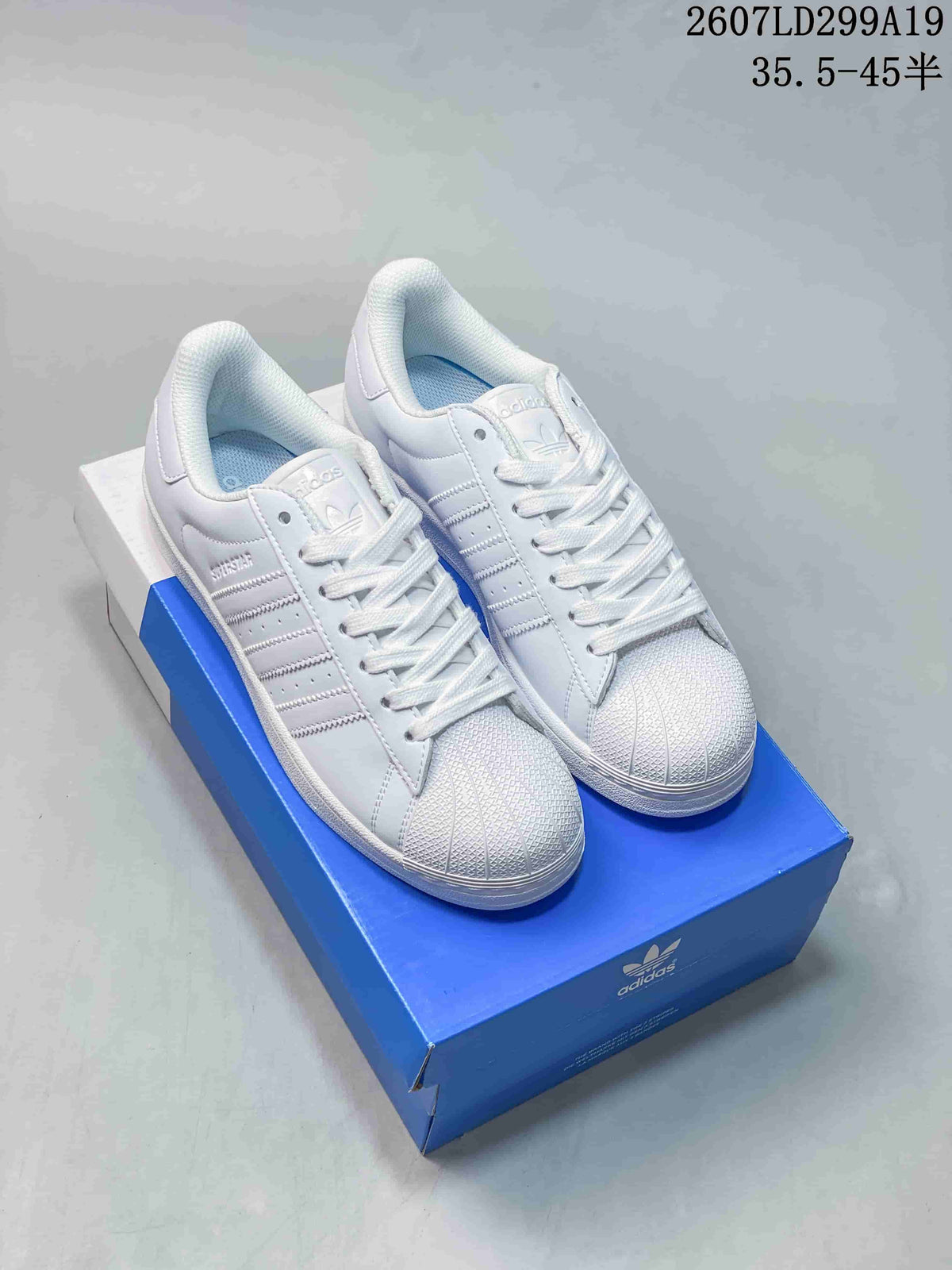 Pair of white Adidas Superstar sneakers displayed on a blue Adidas shoe box, showing the shell toes and lace-up fronts.