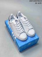 Pair of white Adidas Stan Smith sneakers displayed on a blue Adidas shoebox, with navy heel accents.