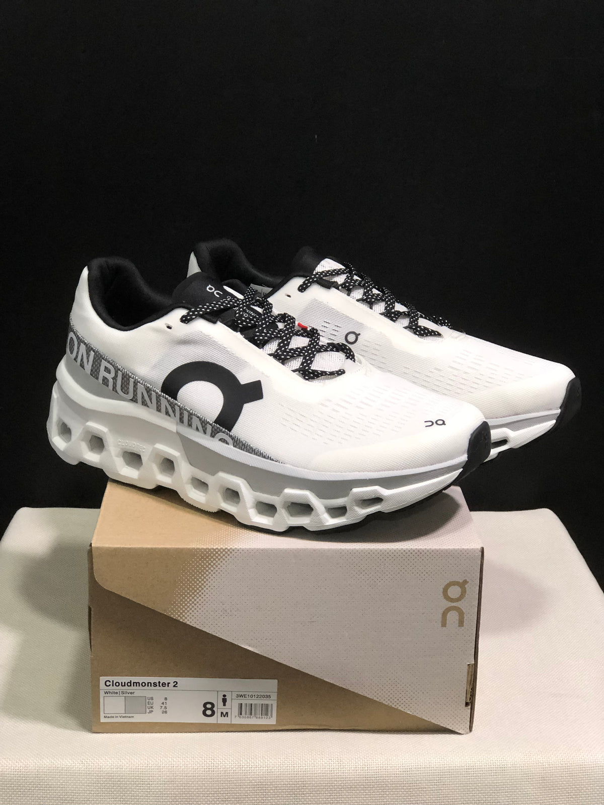 White and silver On Cloudmonster 2 shoes displayed on a box with a visible size label