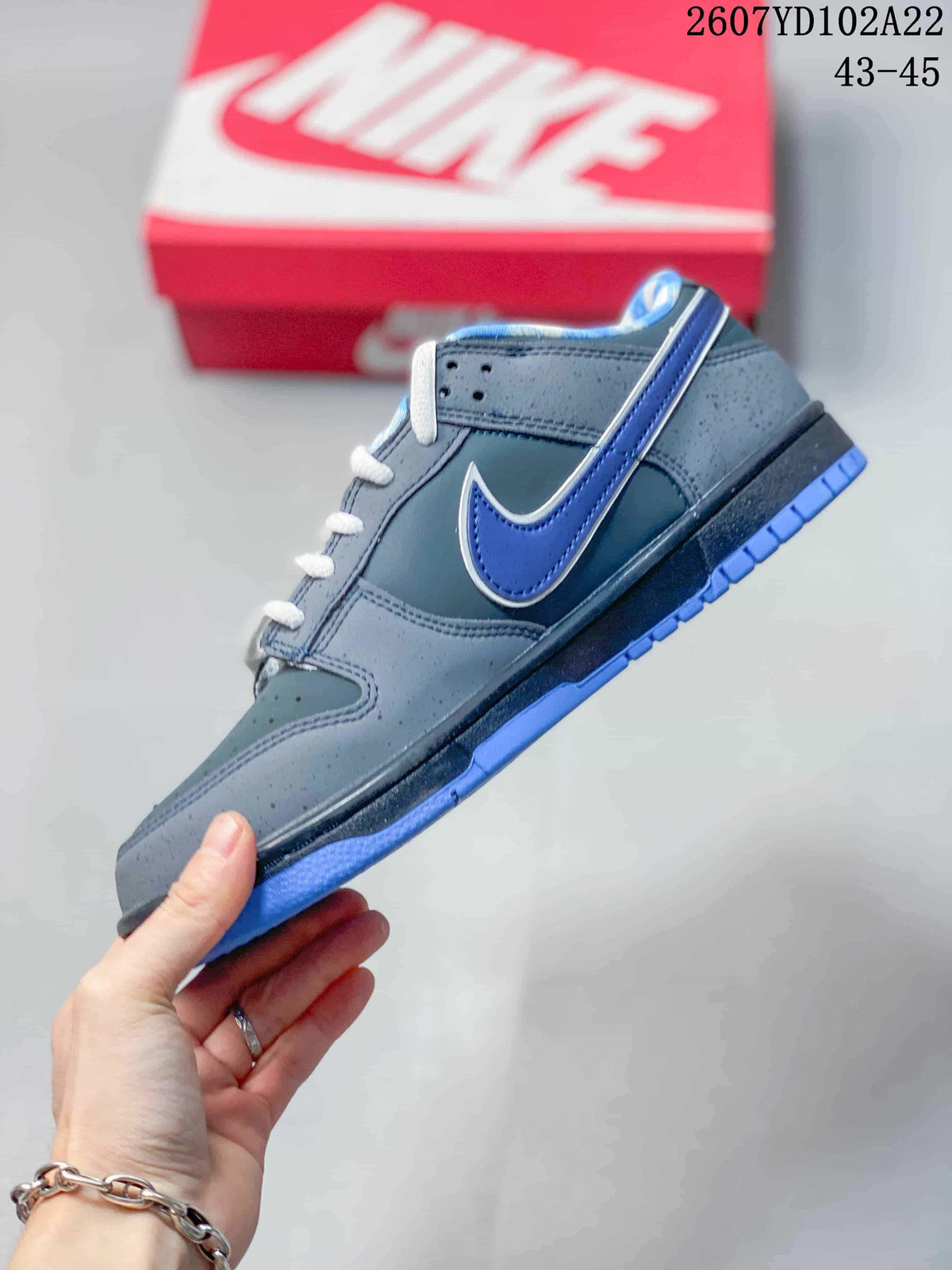 Side view of a grey and blue low-top sneaker showing the contrasting blue side logo, perforated upper panels, and blue outsole.