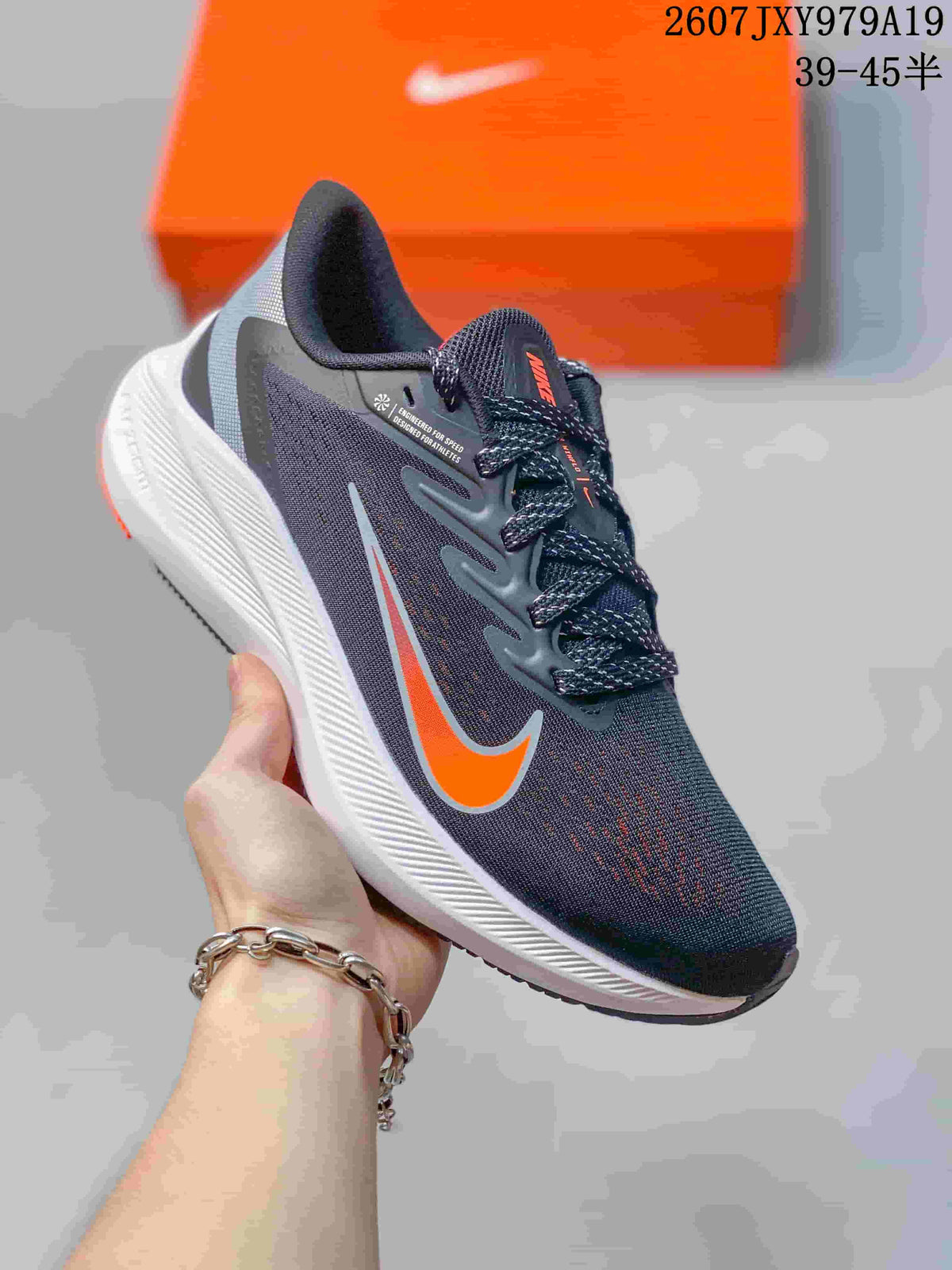 Angled side view of the Nike Zoom Winflo 7 running shoe showing the dark gray upper, orange swoosh, white midsole, and black outsole; image text shows style code 2607JXY979A19 and size range 39–45.