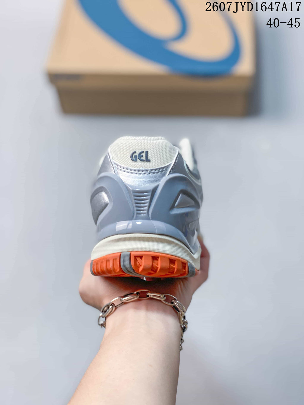 Rear view of an ASICS tennis shoe showing the GEL label, grey heel structure and orange outsole; style code 2607JYD1647A17 and size range 40–45 are visible.