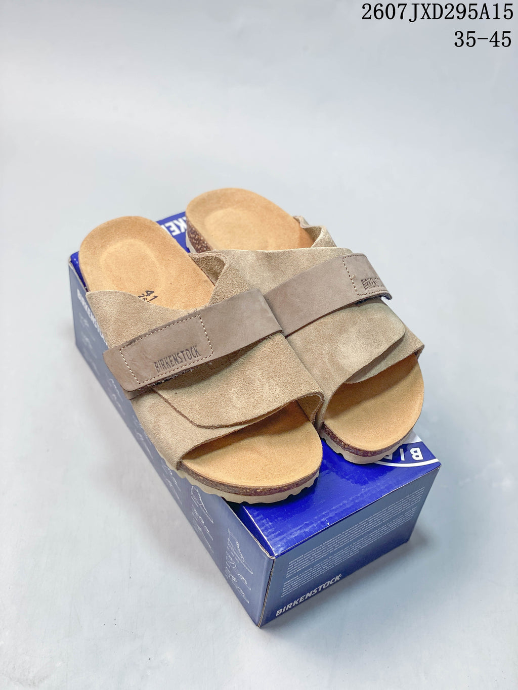 Pair of taupe Birkenstock-style buckle sandals displayed on a blue shoebox; the image shows style code 2607JXD295A15 and size range 35–45.
