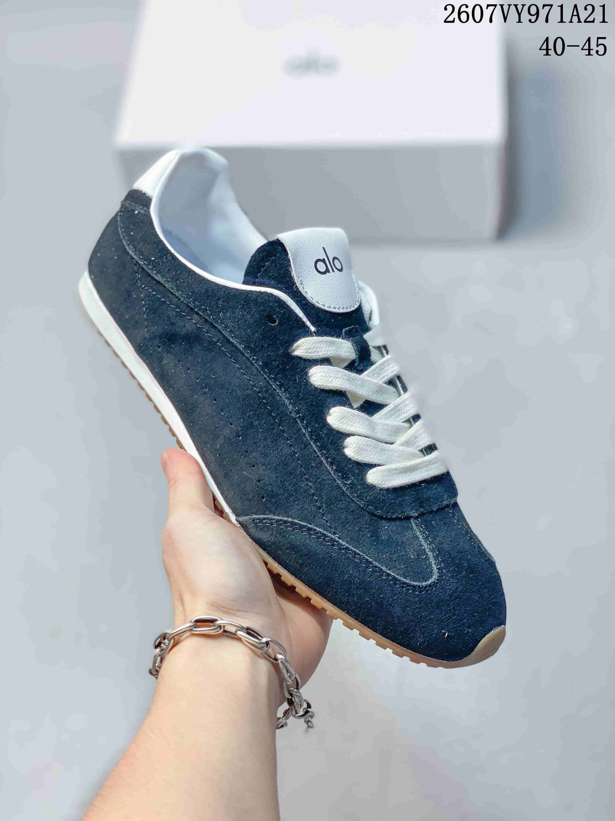 Close side view of a navy Alo Yoga sneaker showing layered panels, perforated detailing, white laces, and a gum-colored outsole.