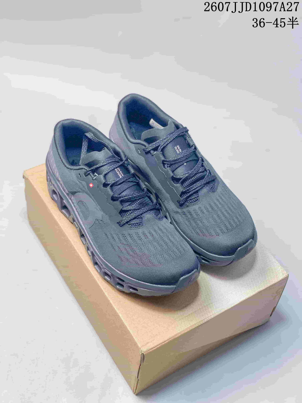 Pair of dark gray On Cloudmonster 3 sneakers displayed on a cardboard box, with the style code and size range visible at the top.
