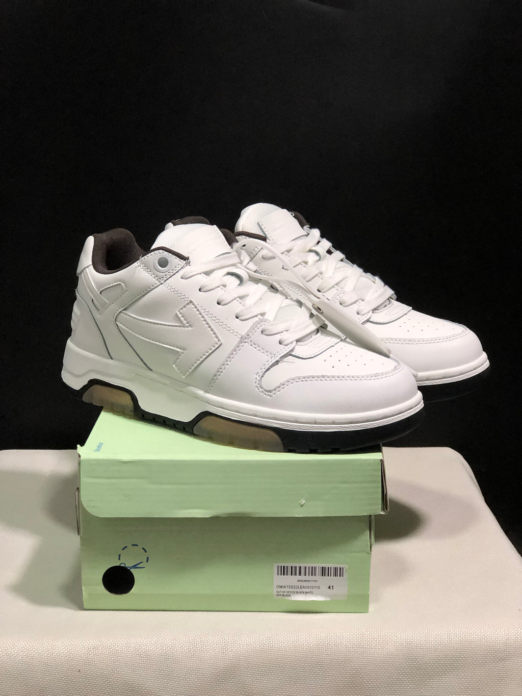 Pair of white low-top sneakers displayed on a pale green shoe box, showing the side panels, laces, and black outsole edges.