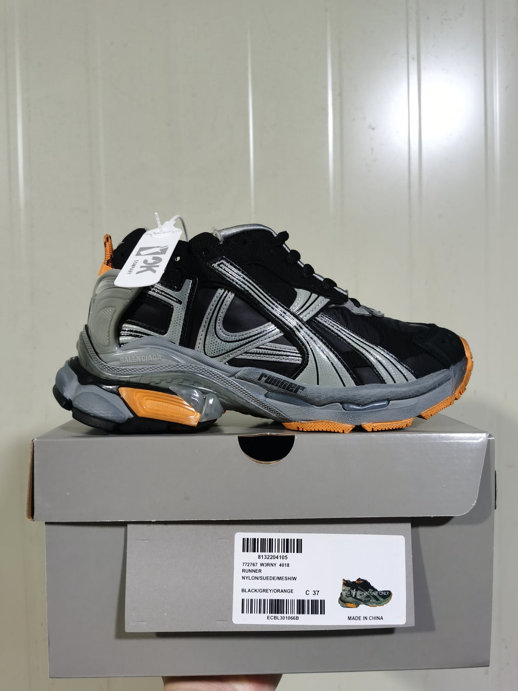 Side view of a black, grey and orange Balenciaga Runner-style sneaker displayed on its box; the box label shows size C 37 and style information.