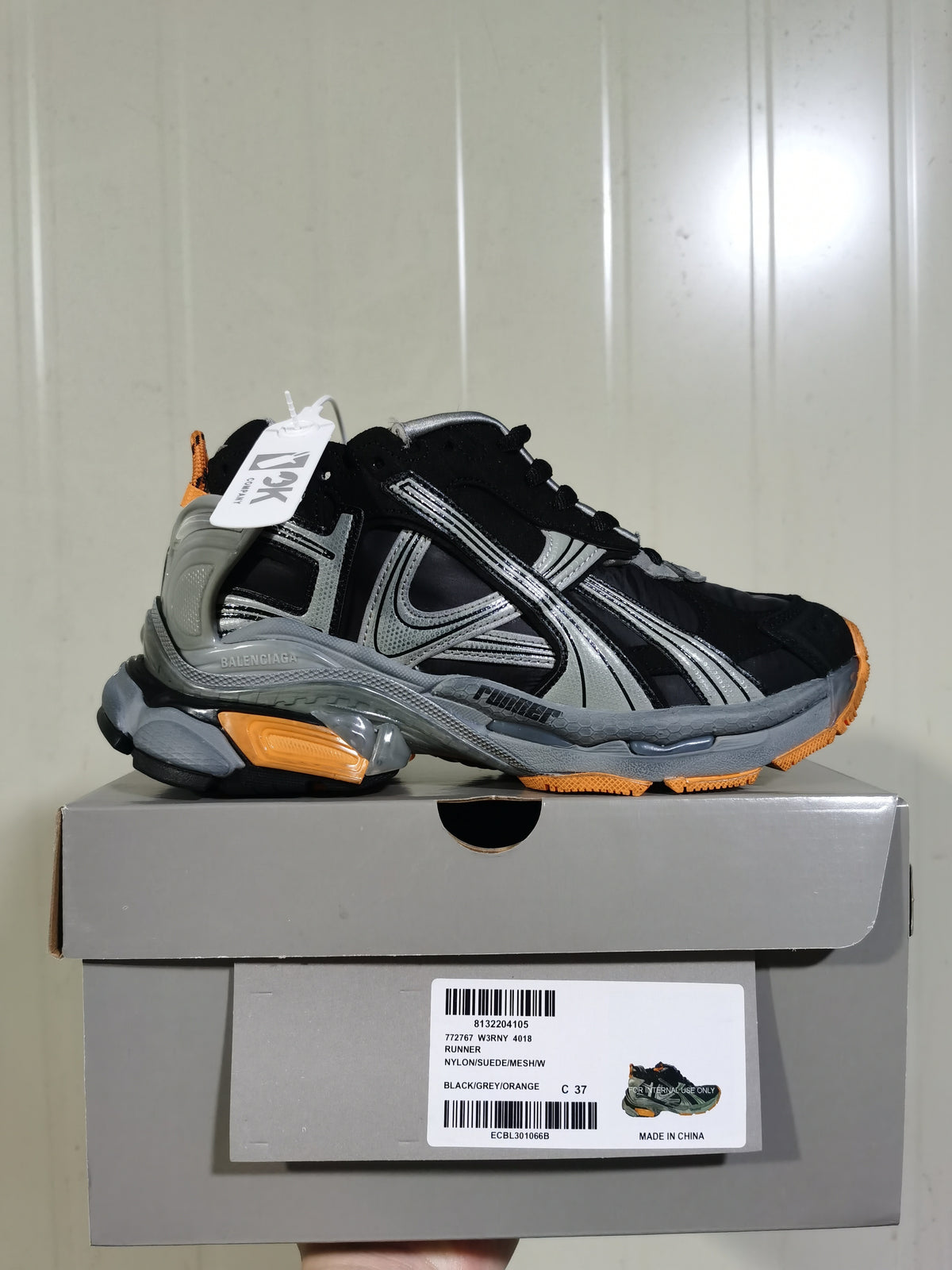 Side view of a black, grey and orange Balenciaga Runner-style sneaker displayed on its box; the box label shows size C 37 and style information.