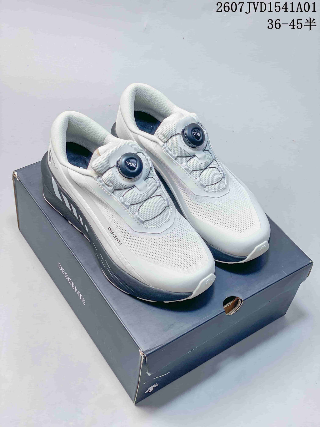 White Descente low-top running shoes with dark gray accents and dial closures, displayed on a box.