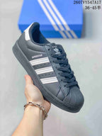 Angled side view of a black Adidas Superstar sneaker showing white stripes, Superstar side branding, and a white heel accent.