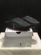 Black Nike Mind 001 slide sandals photographed on a retail box, showing orange outsole pods and side branding.