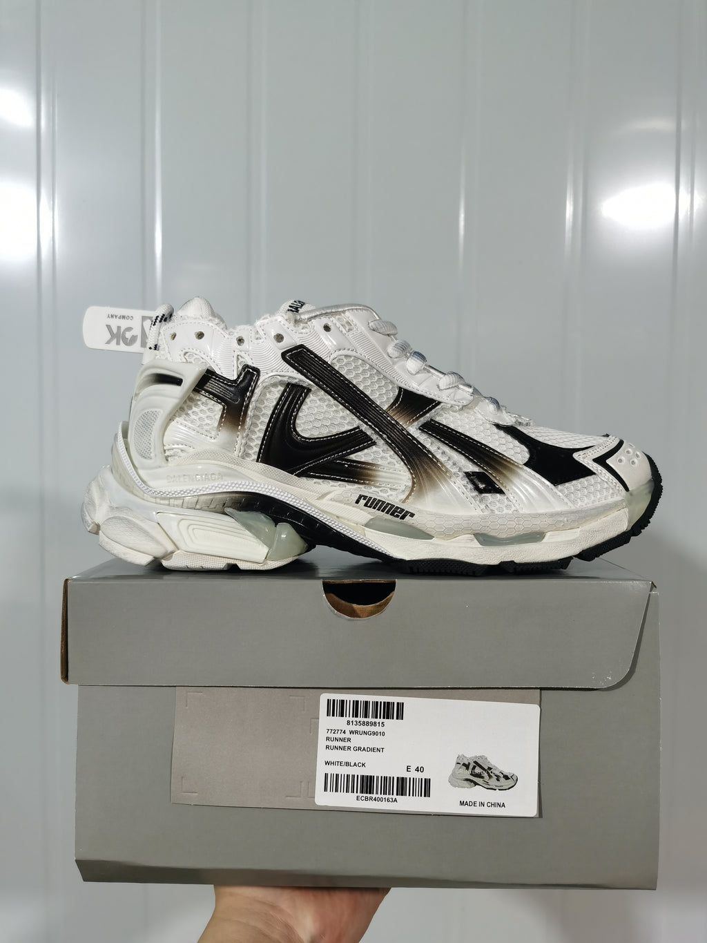 Side-profile product view of the sneaker positioned on its box; the box label shows Runner 9010, White/Black, and size E 40.