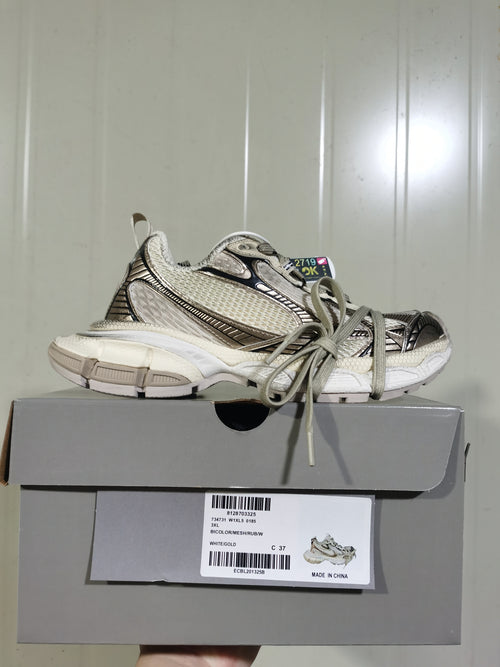 Side view of the white and gold sneaker placed on its box; the box label shows style code 0185 and WHITE/GOLD.