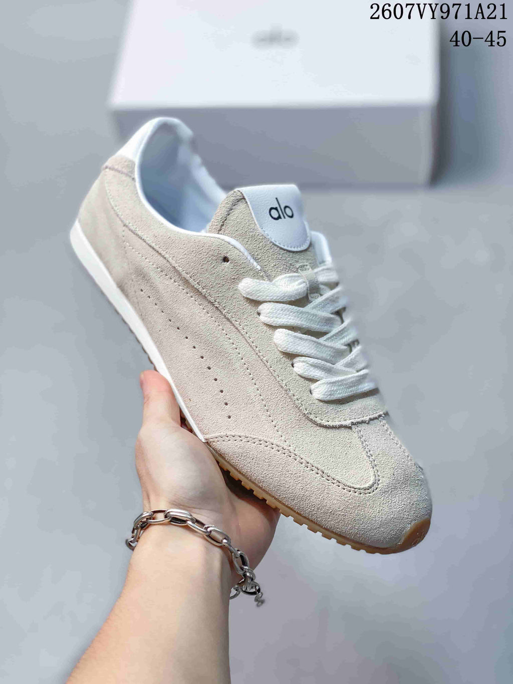 Close side view of a light beige Alo Yoga sneaker showing layered panels, perforated detailing, white laces, and a gum-colored outsole.